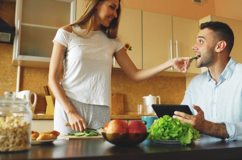 Attractive couple chatting in the kitchen early morning. Beautiful woman feed her husband while cooking breakfast at home